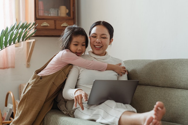 Image of a mother sitting on a couch using a computer while being hugged by her daughter