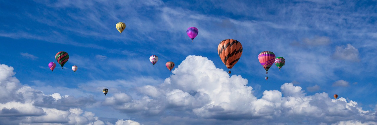 Many colourful hot air balloons float through the blue sky, surrounded by fluffy white clouds. 