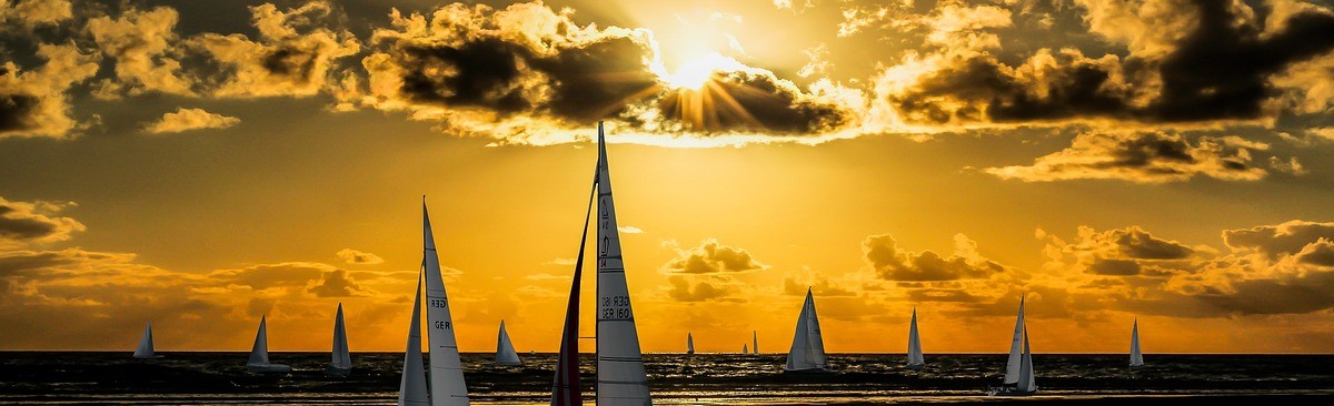 Sailboats on the ocean with a sunset in the background that projects an orange glow across the sky and clouds