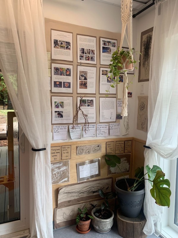 The corner of an ECE centre with a wall of art displayed between two windows with white curtains. 