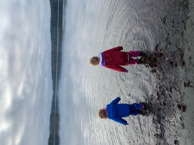 Two children stand in the wet sand at a beach, seen from behind in rain coats on an overcast day